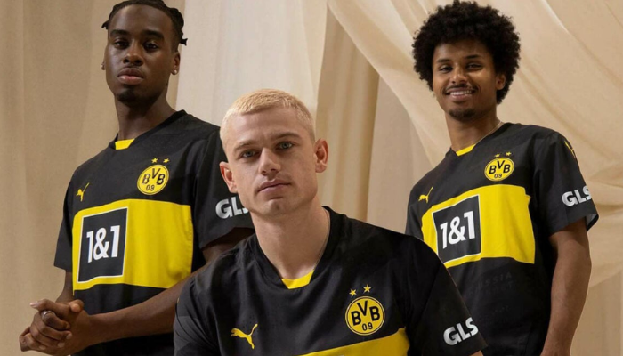 camisa reserva do Borussia Dortmund para a temporada 2024-2025. A peça possui o preto como cor predominante, com detalhes em cinza escuro e amarelo. A frase "United by Borussia" está escrita no interior da camisa em diferentes linguagens, incluindo Braille